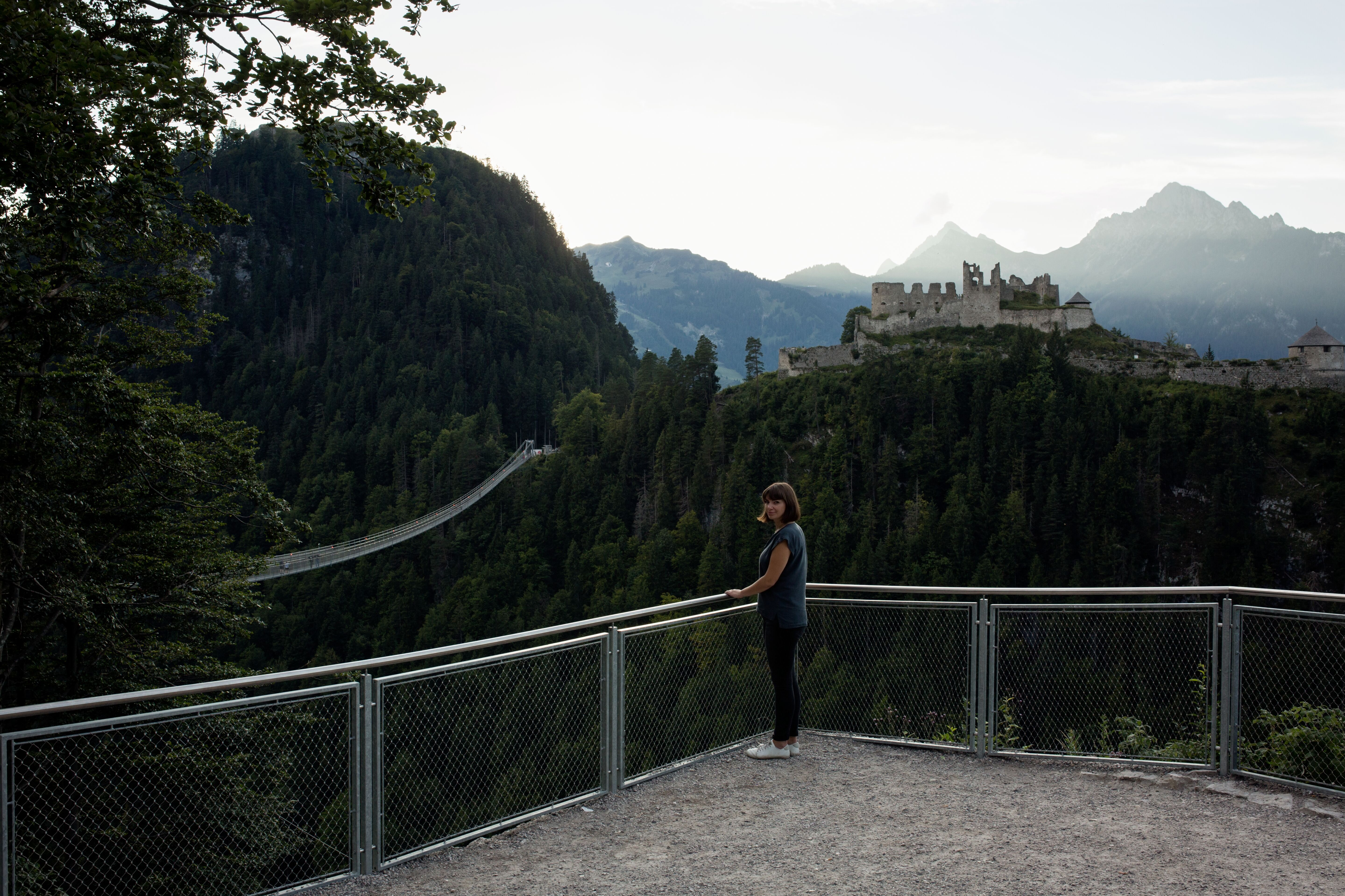 Eine Frau auf einer Aussichtsplattform im Vordergrund, im Hintergrund eine Hängebrücke und eine Festung auf einem Hügel, dahinter hohe Berge