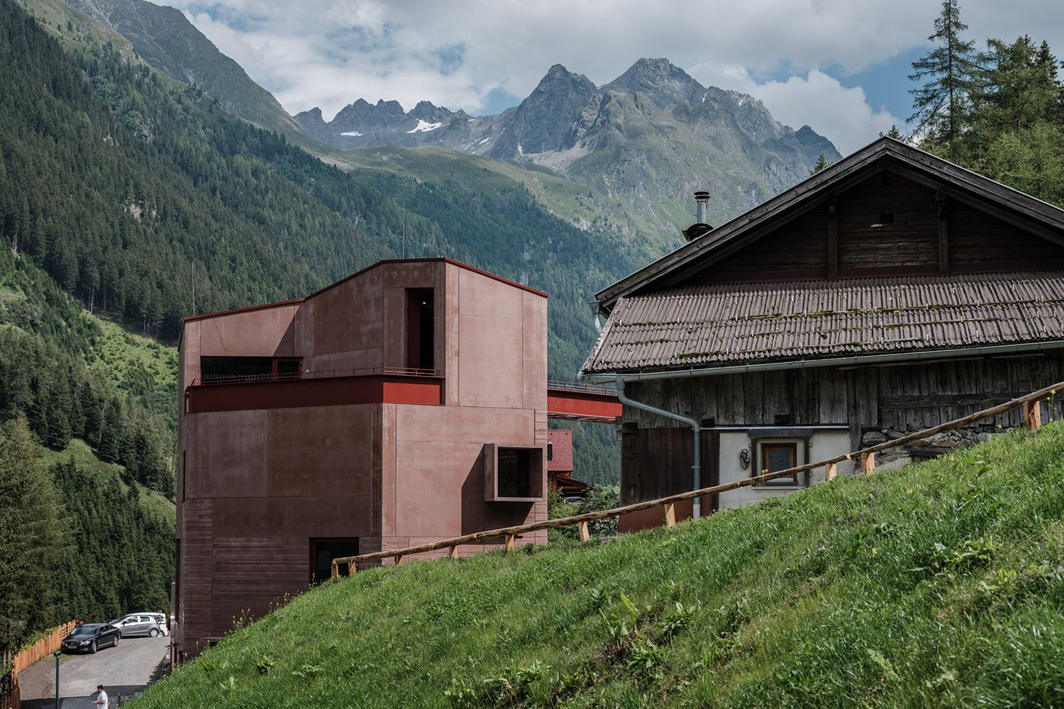 Modernes, rostfarbenes Gebäude, das Steinbockzentrum, steht neben einem traditionellen Wohnhaus im Pitztal, im Hintergrund schroffe Berge